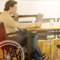 Das Bild zegt einen Mann im Rollstuhl, der an einem Tisch sitzt und sich am PC über eine Berufsunfähigkeitsversicherung informiert.